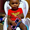Tianna is registered to the contest to win money with this photo: toddler, child, baby_seat, remote_control, red_shirt, wonder_woman_logo, hair_puffs, beads, indoor, carpet, wooden_cabinet, curious, sitting, toy, cute, young_child, person, clothing, footwear_in_background, room
