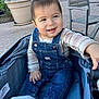 baby, child, denim_overalls, plaid_shirt, stroller, outdoor, smiling, happy, socks, hand, face, person, seated, curious, pavement, chair, planter, plant, daylight, casual_clothing