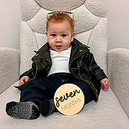 Liam joined the competition — help win amazing prizes! baby, child, leather_jacket, jeans, socks, sunglasses, wooden_sign, plush_chair, indoor, portrait, cute, infant, smiling, seated, clothing, furniture, young_child, happy, person, home