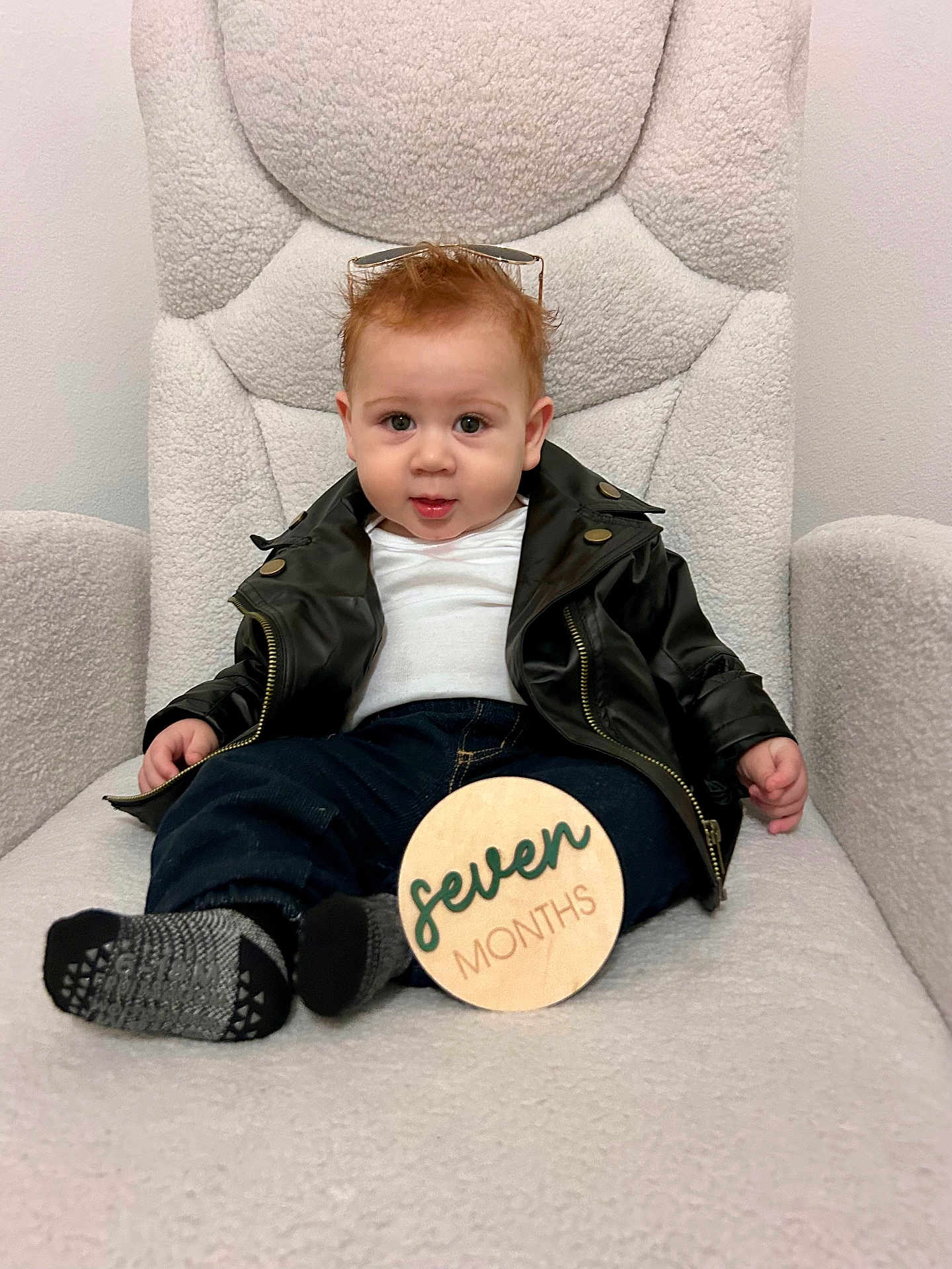 Liam joined the competition — help win amazing prizes! baby, child, leather_jacket, jeans, socks, sunglasses, wooden_sign, plush_chair, indoor, portrait, cute, infant, smiling, seated, clothing, furniture, young_child, happy, person, home