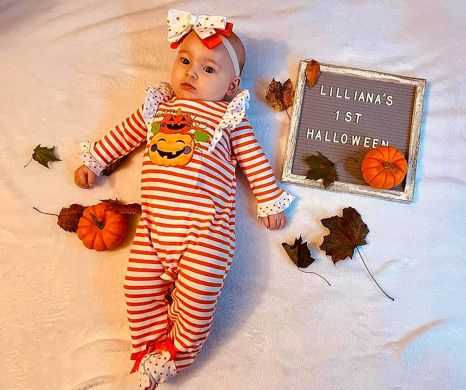 Lilliana is registered to the contest to win money with this photo: baby_toddler_clothing, baby_toys, brown, calabaza, child, costume, doll, fashion_accessory, font, happy, headwear, orange, pattern, people_in_nature, person, personal_protective_equipment, product, pumpkin, sleeve, stuffed_toy