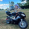 Luis is registered to the contest to win money with this photo: toddler, child, motorcycle, toy, grass, outdoor, blue_sky, clouds, cap, jacket, vehicle, parked_truck, trees, road, daylight, happy, smiling, playtime, nature, recreation
