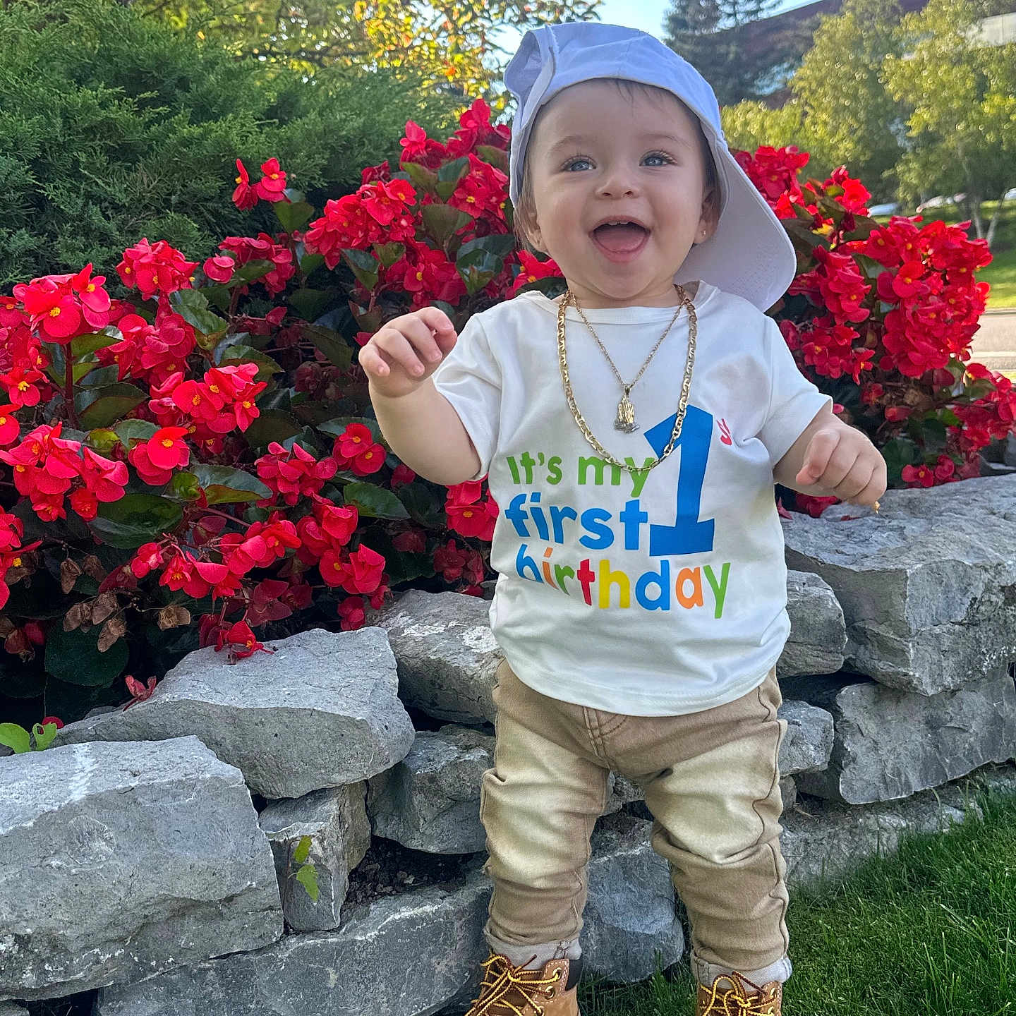 Luis joined the competition — help win amazing prizes! toddler, child, birthday, first_birthday, outdoor, flowers, red_flowers, grass, stone_wall, hat, cap, boots, pants, tshirt, gold_chain, smiling, happy, nature, daylight, person