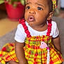 baby, toddler, child, headband, bow, plaid_dress, colorful_clothing, pacifier_clip, sitting, indoors, potted_plant, blurred_background, rug, bare_feet, big_eyes, surprised_expression, portrait, candid, close_up, adorable