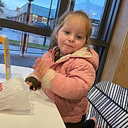 Bonnie joined the competition — help win amazing prizes! buildings, casual_clothing, chair, child, curly_hair, daylight, expression, face, hand_movement, indoor, leaf_pattern, person, pink_jacket, playful, seated, smiling, streetlamp, table, window, young_child