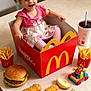 toddler, child, happy_meal, mcdonalds, fries, burger, chicken_nuggets, apple_slices, soft_drink, toy_car, pink_dress, table, smiling, happy, food, fast_food, restaurant, indoor, cute, playful
