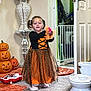 child, girl, halloween_costume, dress, indoor, carpet, pumpkin_decoration, toy, door, vase, flowers, baby_gates, socks, orange, black, white, home, floor, decor, playful
