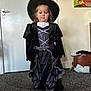 child, witch_costume, black_hat, black_dress, indoor, carpet, door, bag, diapers, portrait, person, wall_art, standing, young_child, costume, dress_up, room, floor, photograph, hat