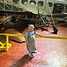 airplane, aviation, backpack, child, concrete_floor, dress, floor, footwear, girl, hangar, indoor, light, metal_structure, pigtails, shadow, smile, transport, vintage_airplane, window, yellow_barrier