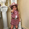 child, cowgirl, cowboy_hat, pink_outfit, western_style, boots, thumbs_up, portrait, smiling, indoors, decorative_column, flower_arrangement, ornate_furniture, carpet, standing, fashion, costume, young_girl, full_body, home_decor
