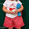 child, girl, smiling, portrait, standing, door, green_door, plush_toy, blue_toy, red_shoes, hair_ribbons, pigtails, white_shirt, red_shorts, checkered_clothing, cherries, container, doormat, outdoor_entryway, joyful