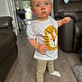 Harry is registered to the contest to win money with this photo: toddler, child, standing, blue_eyes, white_tshirt, lion_graphic, beige_pants, socks, wooden_floor, living_room, leather_couch, furniture, interior, doorway, modern_home, person, cute, indoor, young_child, portrait