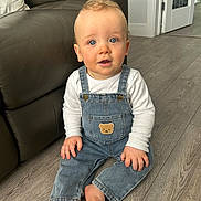 Harry joined the competition — help win amazing prizes! baby, child, denim_overalls, white_shirt, blue_eyes, curly_hair, indoor, wooden_floor, leather_couch, teddy_bear_patch, barefoot, sitting, cute, portrait, home, curious_expression, furniture, flooring, person, infant