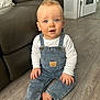baby, child, denim_overalls, white_shirt, blue_eyes, curly_hair, indoor, wooden_floor, leather_couch, teddy_bear_patch, barefoot, sitting, cute, portrait, home, curious_expression, furniture, flooring, person, infant