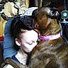 affectionate, animal, black_fur, brindle_dog, brown_fur, closeup, companion, cuddling, dog, fan, furniture, home, hugging, indoors, large_dog, person, pet, pink_collar, portrait, seated