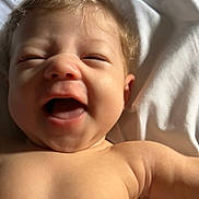 Ava is registered to the contest to win money with this photo: baby, laughing, smiling, skin, hair, face, child, infant, bare_chest, arm, white_sheet, sunlight, happy, closeup, portrait, cute, joy, lying_down, expression, natural_light