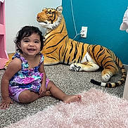 Jasmine joined the competition — help win amazing prizes! child, toddler, smiling, sitting, plush_toy, tiger, carpet, indoor, colorful_clothing, furniture, wall, pink, blue, toy, cute, happy, room, hair, person, floor
