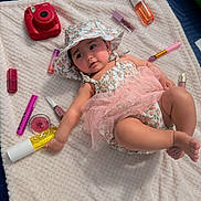 Layla is registered to the contest to win money with this photo: baby, infant, child, portrait, lying_down, floral_hat, pink_dress, tulle_skirt, makeup, cosmetics, makeup_brushes, lipstick, lip_gloss, skincare, instant_camera, blanket, indoor, top_down_view, smiling, fashion