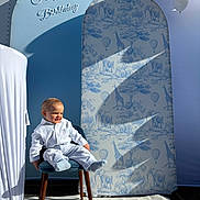 Noé participe au concours pour gagner de l'argent avec cette photo : child, toddler, boy, smile, birthday_backdrop, chair, balloons, sunlight, shadow, patterned_screen, elephant_pattern, giraffe_pattern, hot_air_balloon_pattern, white_shirt, socks, portrait, celebration, wooden_chair_legs, outdoor, fabric