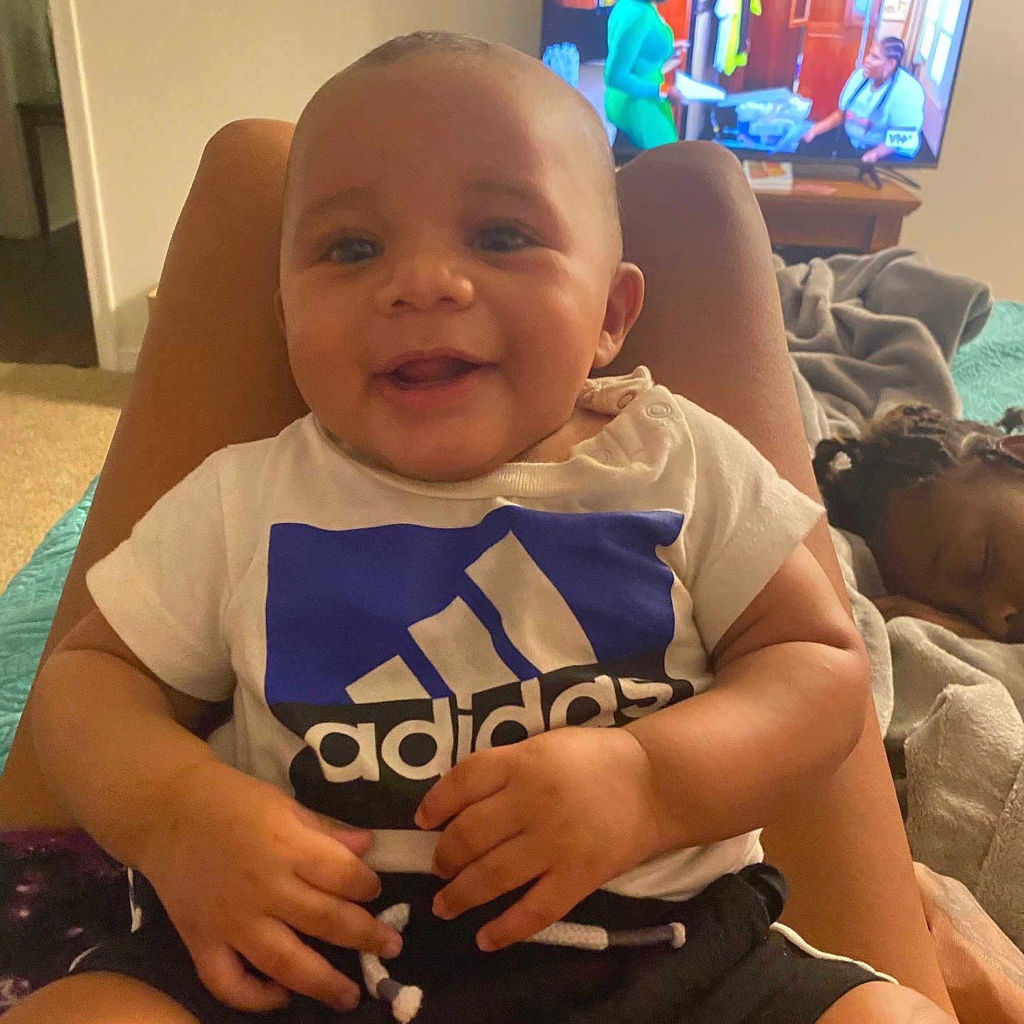 Kiair is registered to the contest to win money with this photo: adidas, baby, bed, blanket, child, cozy, furniture, hair, hands, happy, indoor, legs, living_room, person, relaxing, shorts, sleeping_child, smiling, television, tshirt