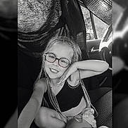 Judith participe au concours pour gagner de l'argent avec cette photo : black_and_white, braids, car_interior, casual_clothing, child, cute, fabric, girl, glasses, hand_on_chin, happy, playful, portrait, seat, seat_cover, sitting, smiling, steering_wheel, window, young