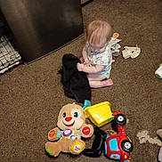 Estella is registered to the contest to win money with this photo: baby, child, carpet, toy, plush_toy, stuffed_animal, floor, indoor, person, sitting, clothing, play, cute, infant, young_child, carpeted_floor, toy_truck, black_cloth, room, home