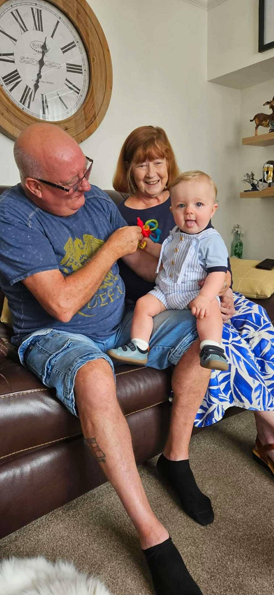 Alfie is registered to the contest to win money with this photo: adult, baby, carpet, casual_clothing, child, clock, decor, family, footwear, glasses, happy, indoor, living_room, people, playful, shelf, smiling, sofa, tattoo, toy