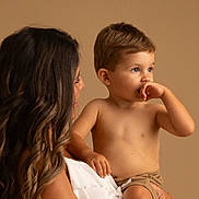 Kélyan a rejoint le concours — aidez-le/la à gagner de superbes lots ! woman, child, toddler, holding, portrait, indoor, studio, brown_background, white_clothing, curly_hair, bare_chest, shorts, cute, family, mother, affection, looking_away, skin, intimate, soft_lighting