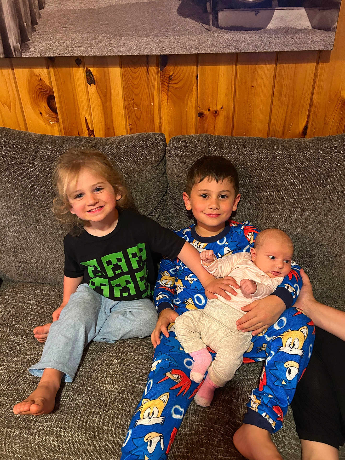 Kash is registered to the contest to win money with this photo: child, baby, pajamas, couch, smile, barefoot, indoor, wood_paneling, family, sibling, holding, happy, clothing, cute, relaxed, portrait, person, infant, casual, seated