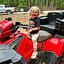 child, blond_hair, curly_hair, atv, red_vehicle, outdoor, woods, truck, tshirt, smiling, crocs, footwear, playful, dirt, grip_handle, seat, nature, daylight, person, vehicle
