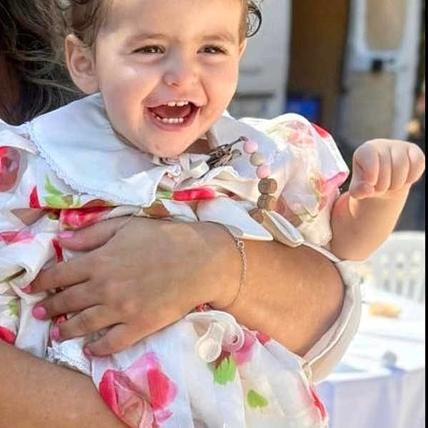 Eliya a rejoint le concours — aidez-le/la à gagner de superbes lots ! adult_hand, chair, child, clothing, curly_hair, daylight, event, face, floral_dress, happy, holding, legs, outdoor, person, smiling, sunlight, table, toddler, trees, white_sandals
