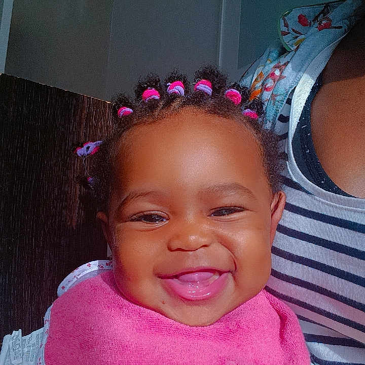 Joyce Ynhelia a rejoint le concours — aidez-le/la à gagner de superbes lots ! apparel, baby, child, closeup, cute, face, hair_ties, happy, human, indoor, infant, joyful, person, pink_bib, portrait, seated, smiling, striped_shirt, yellow_clothing, young_child