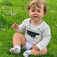 Tiana a rejoint le concours — aidez-le/la à gagner de superbes lots ! toddler, child, grass, outdoor, greenery, flower, white_shirt, socks, curly_hair, cute, baby, nature, sitting, smile, person, daylight, lawn, young_child, casual_clothing, playful