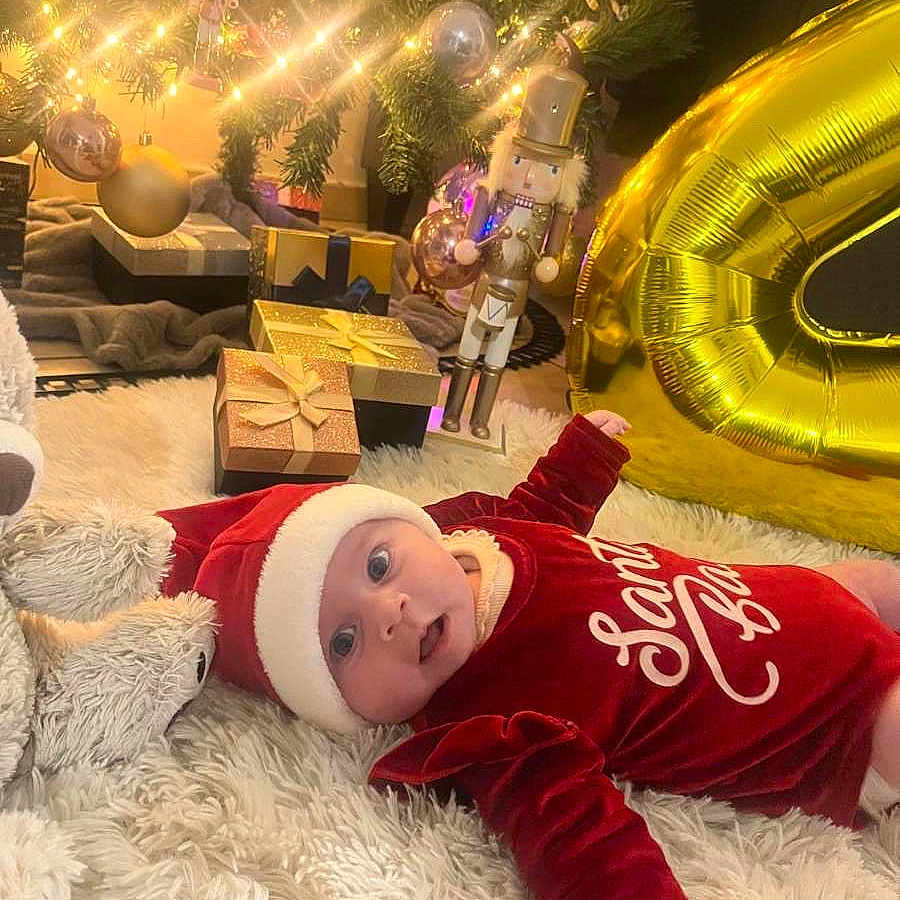 Milya a rejoint le concours — aidez-le/la à gagner de superbes lots ! baby, celebration, christmas_tree, cute, decorations, festive, fluffy_rug, gold_balloon, holiday, holiday_season, indoors, lights, nutcracker, ornaments, presents, red_clothing, santa_hat, smiling, toy, winter