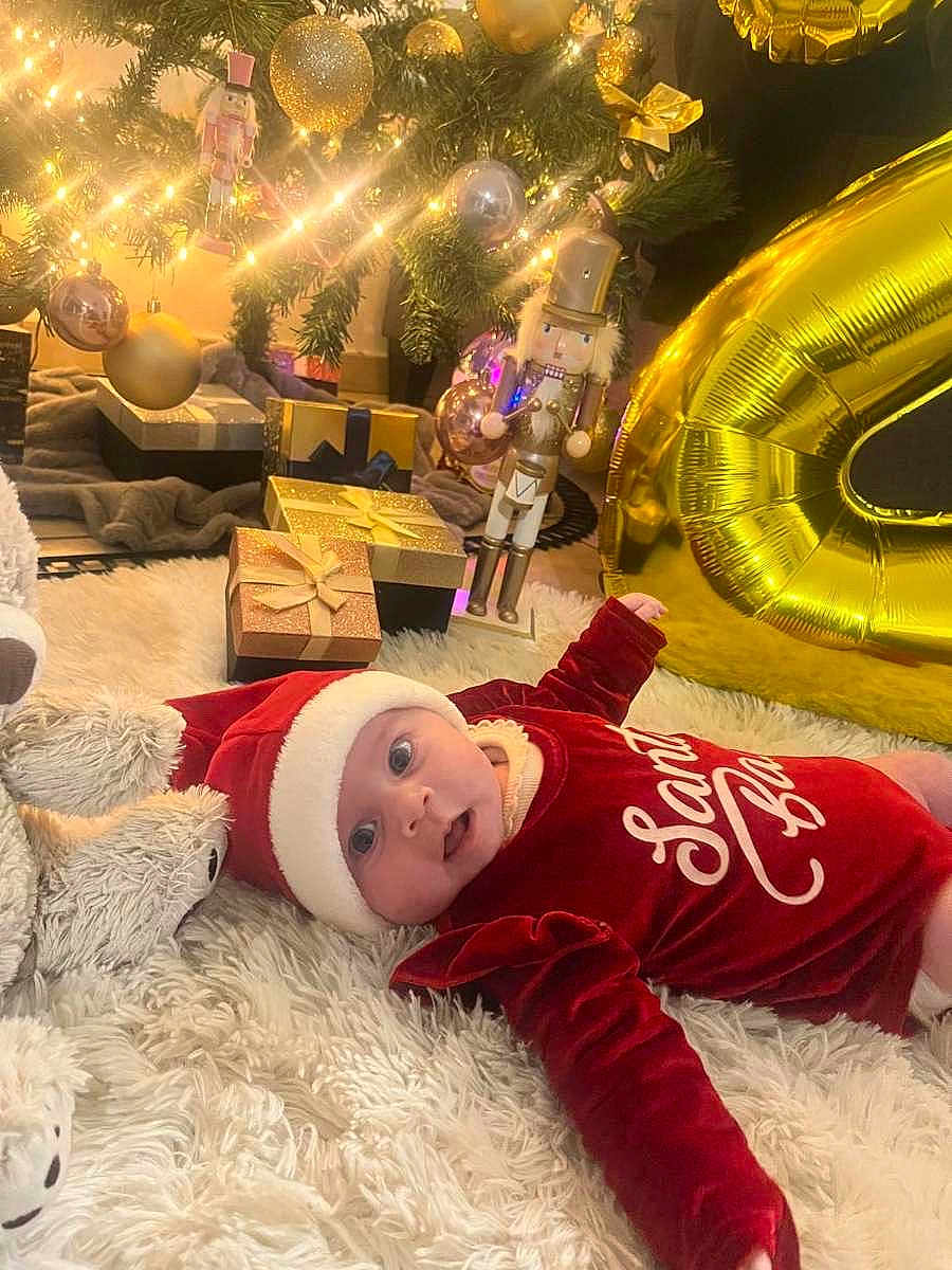 Milya a rejoint le concours — aidez-le/la à gagner de superbes lots ! baby, christmas_tree, holiday, presents, ornaments, lights, santa_hat, red_clothing, fluffy_rug, toy, gold_balloon, nutcracker, festive, celebration, indoors, decorations, smiling, cute, winter, holiday_season