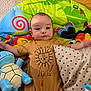baby, child, plush_toy, stuffed_animal, play_mat, pillow, polka_dot, tshirt, lion_graphic, floor, wooden_floor, colorful, smiling, infant, cute, indoors, toy, person, young_child, lying_down