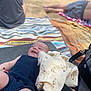 baby, smiling, blanket, beach, sand, cloth, person, infant, outdoor, relaxing, summer, happy, lying_down, colorful, pattern, people, casual, sunny, vacation, family