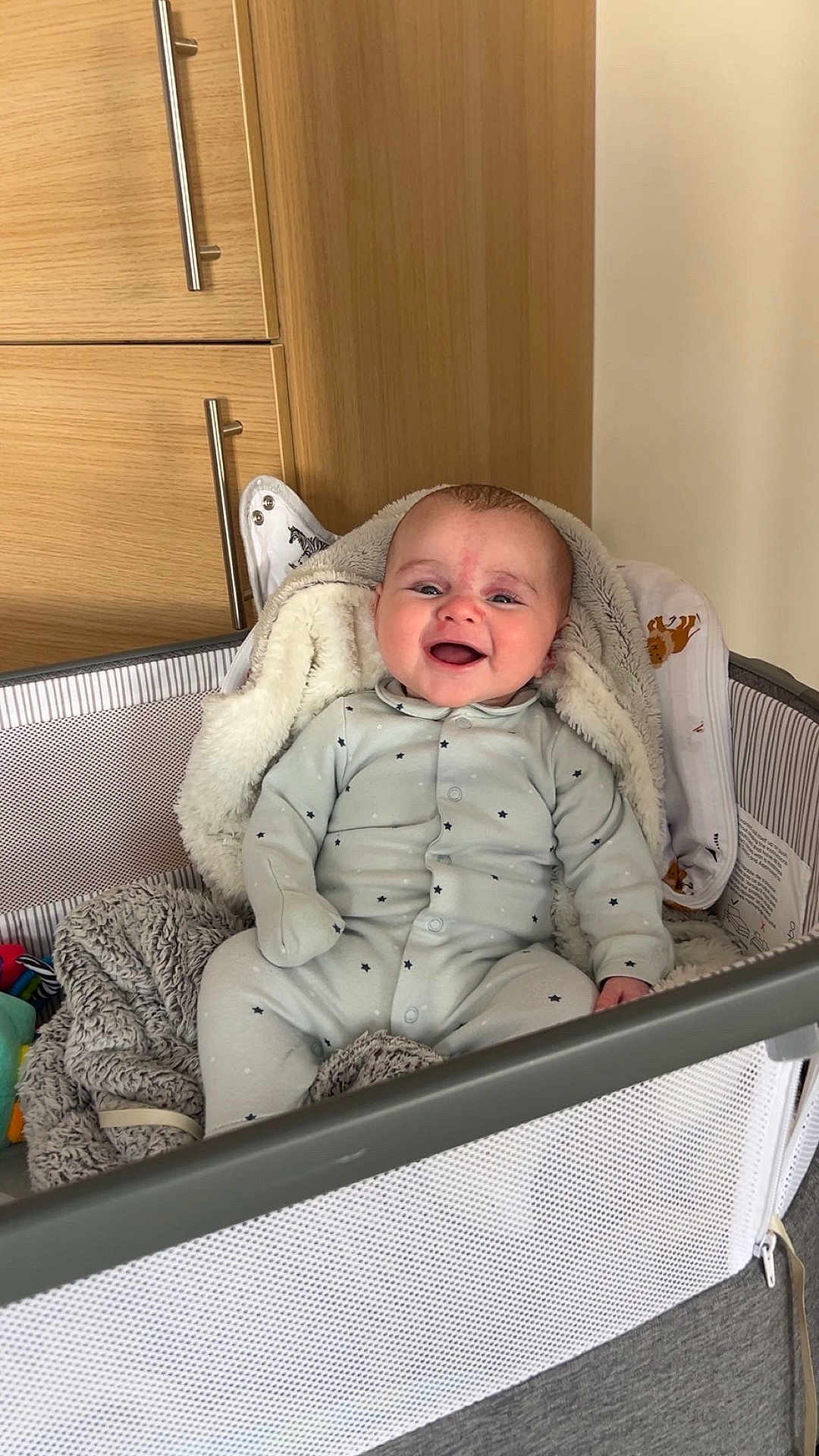 Oscar is registered to the contest to win money with this photo: baby, infant, smiling, playpen, blanket, onesie, cozy, indoor, furniture, child, happy, cute, soft, toy, fabric, head, face, sitting, room, wooden_cabinet