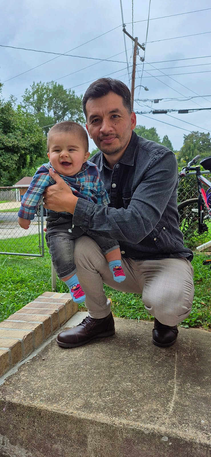 Esteban Muñoz joined the competition — help win amazing prizes! baby, child, man, outdoor, smiling, plaid_shirt, denim_jacket, grass, bike, fence, sidewalk, socks, elmo, casual_clothing, portrait, family, happy, tree, person, daylight
