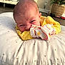 baby, infant, pillow, yellow_clothing, face, expression, home_interior, floor, blanket, soft_texture, newborn, person, cute, lying_down, indoor, decor, pattern, closeup, head, sleeve