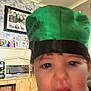 child, face, green_hat, indoor, living_room, furniture, photograph, expression, person, cabinet, decor, portrait, blurry, mouth_open, headwear, curly_hair, wall_art, shelf, household, casual