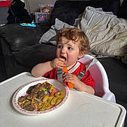 Riley joined the competition — help win amazing prizes! toddler, child, high_chair, eating, food, plate, fork, curly_hair, red_shirt, living_room, couch, pillow, table, indoor, meal, cute, person, baby, snack, happy