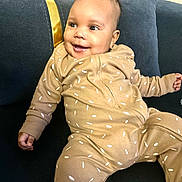 Caëlis-Aïyann participe au concours pour gagner de l'argent avec cette photo : baby, infant, smiling, onesie, brown_clothing, sprinkles_pattern, couch, dark_blue_couch, pillow, indoor, happy, cute, child, person, portrait, seated, living_room, cozy, soft_lighting, relaxed