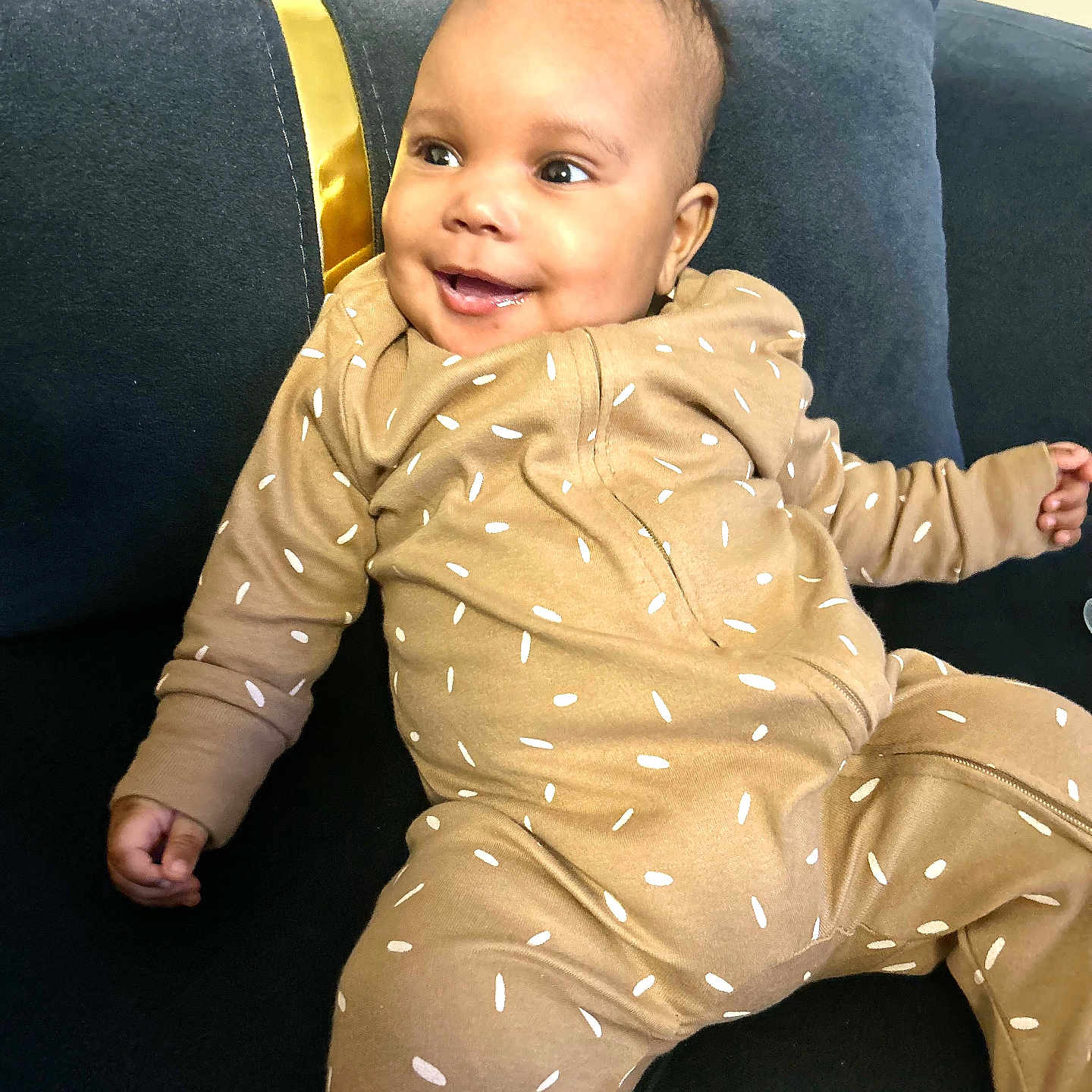 Caëlis-Aïyann participe au concours pour gagner de l'argent avec cette photo : baby, brown_clothing, child, couch, cozy, cute, dark_blue_couch, happy, indoor, infant, living_room, onesie, person, pillow, portrait, relaxed, seated, smiling, soft_lighting, sprinkles_pattern