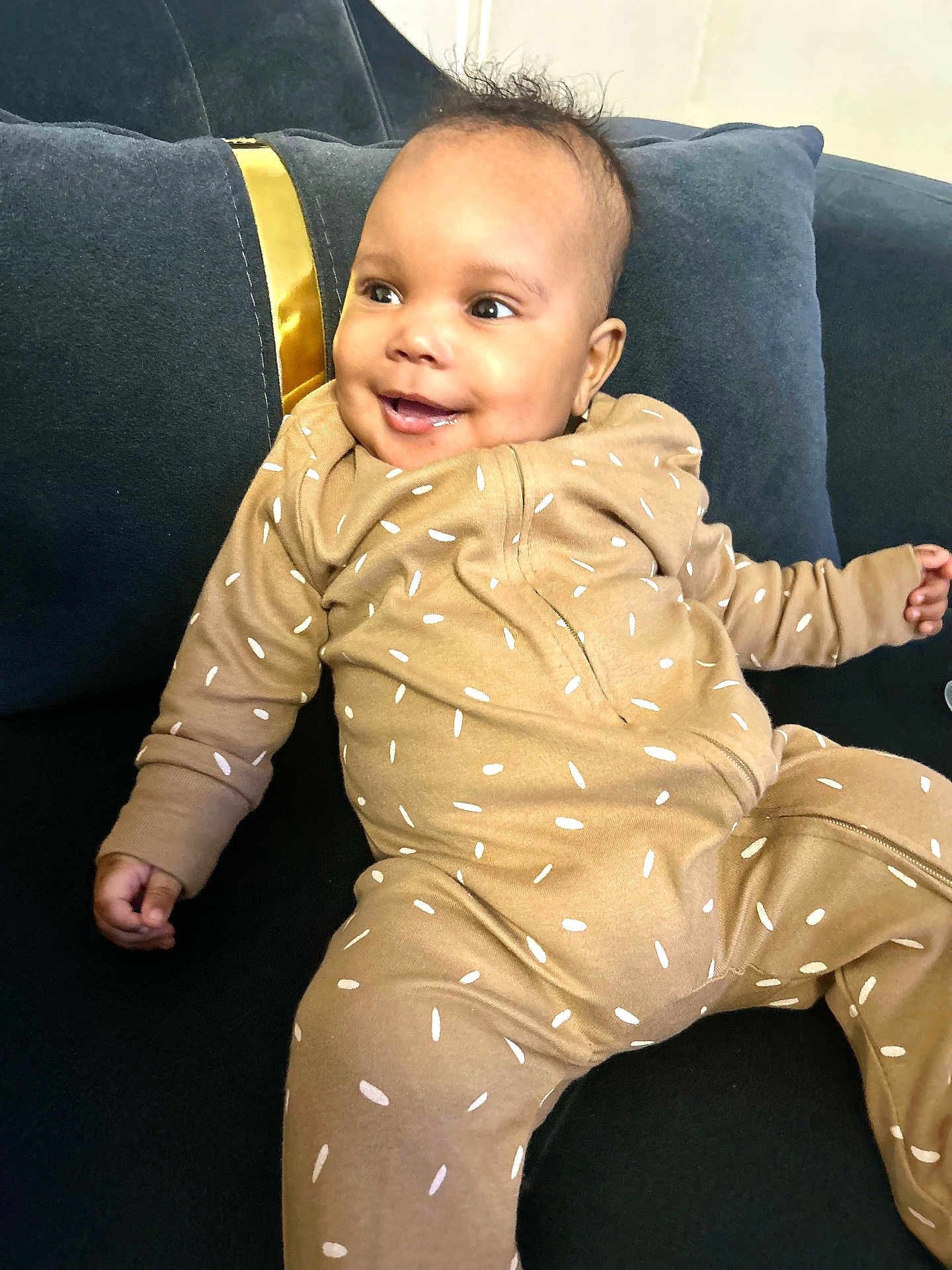 Caëlis-Aïyann participe au concours pour gagner de l'argent avec cette photo : baby, infant, smiling, onesie, brown_clothing, sprinkles_pattern, couch, dark_blue_couch, pillow, indoor, happy, cute, child, person, portrait, seated, living_room, cozy, soft_lighting, relaxed