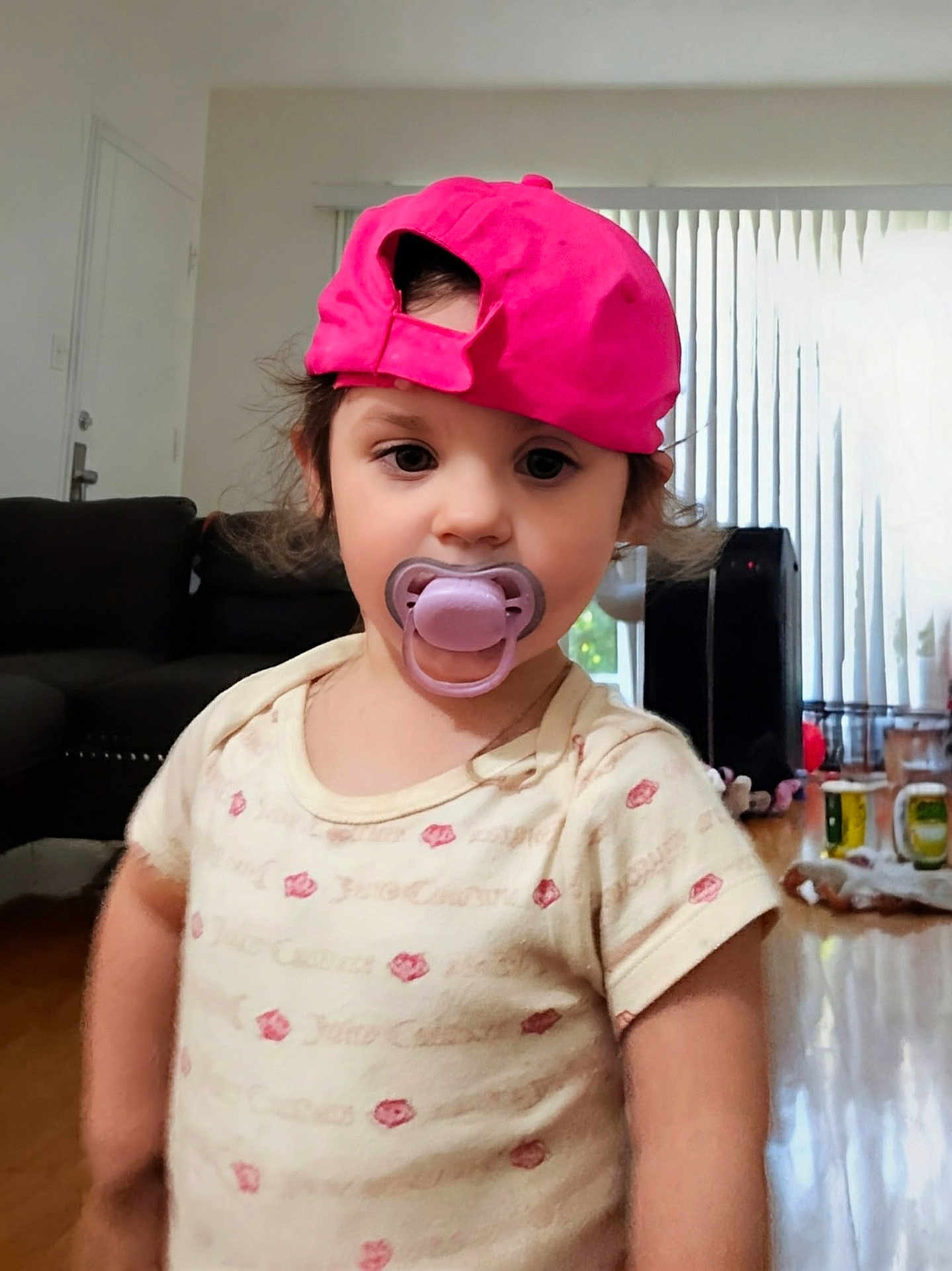 Cersei joined the competition — help win amazing prizes! toddler, child, pacifier, pink_cap, indoor, living_room, curtains, couch, wooden_floor, short_sleeve_shirt, person, young_child, face, hair, expressionless, standing, home, window, furniture, daylight