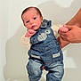 baby, infant, denim_overalls, cute_clothing, white_shirt, baby_shoes, mickey_mouse, standing, adult_hands, indoor, portrait, child, small_fist, denim, blue_clothing, footwear, person, studio_lighting, soft_background, expression