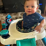 Jaylin is registered to the contest to win money with this photo: baby, child, toddler, smiling, portrait, indoor, walker, baby_walker, toy, colorful_toy, blue_shirt, shark_pattern, seated, face, brown_hair, home, living_room, carpet, furniture, happy