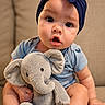 baby, infant, plush_toy, elephant, stuffed_animal, headband, blue_clothing, sitting, couch, beige_background, cute, child, indoors, soft_toy, facial_expression, portrait, small_child, home, comfort, playful