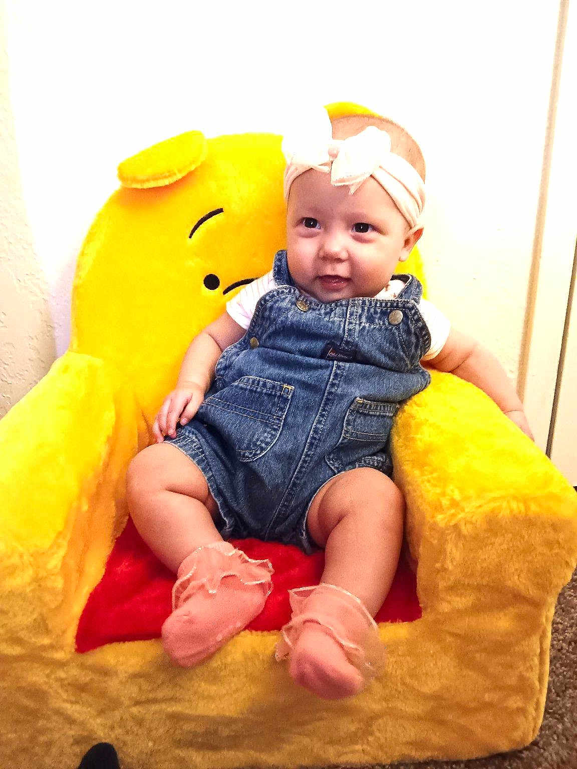 Hazel joined the competition — help win amazing prizes! baby, child, denim_overalls, headband, pink_socks, plush_chair, yellow_chair, smiling, indoor, cute, happy, infant, seated, soft_toys, cozy, portrait, young_child, furniture, childhood, comfortable